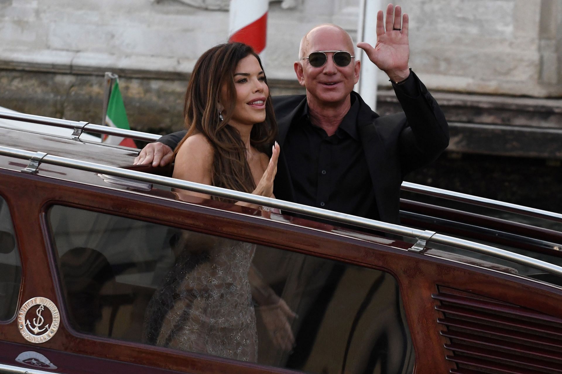 Jeff Bezos and Lauren Sanchez wave as they exit the Hotel Aman - (Image via Getty)